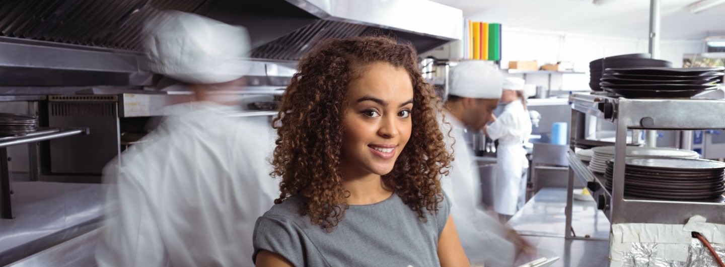 Junge Frau in Businesslook steht zwischen arbeitenden Köchen in Gastroküche