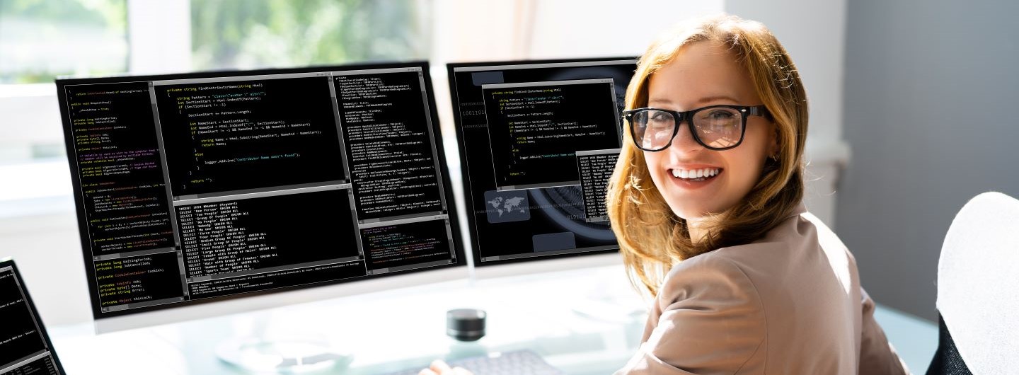 PHP-Programmierer Weiterbildung: Eine junge Programmiererin mit schwarzer Brille sitzt am PC mit zwei Monitoren und lächelt in die Kamera