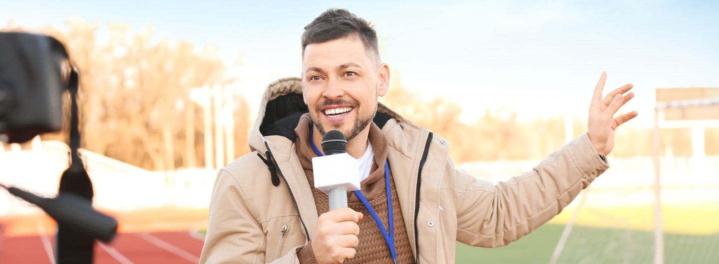 Sportjournalist moderiert lachend in Kamera vor Sportplatz